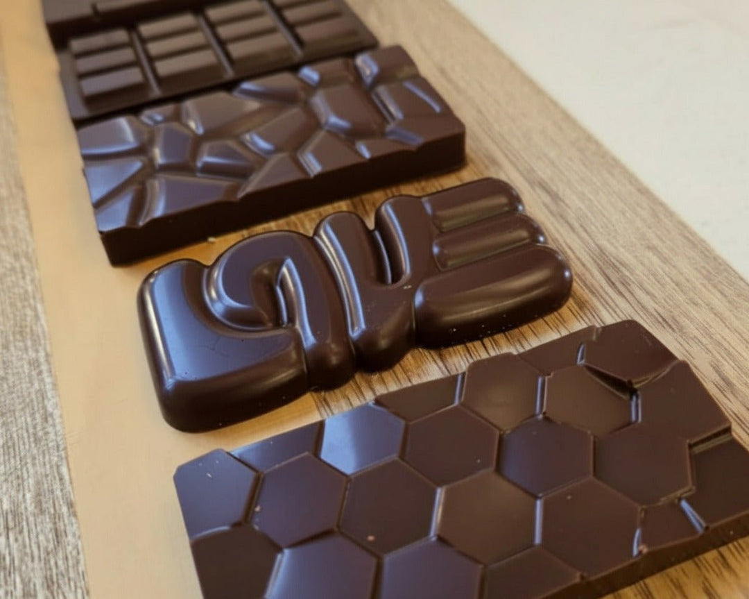 Four dark chocolate bars with different textured patterns, including a honeycomb design, arranged diagonally on a light-colored wooden board.