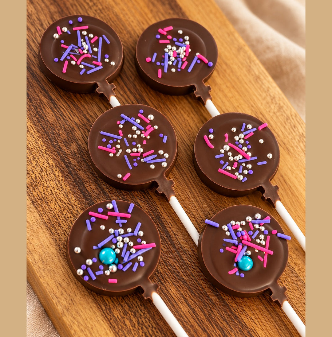 Six round chocolate lollies decorated with pink, purple and silver sprinkles on a wooden board.